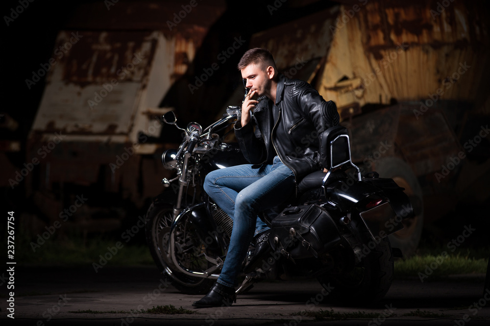 Cute biker in leather jacket sits on a motorcycle. Smoking a cigarette