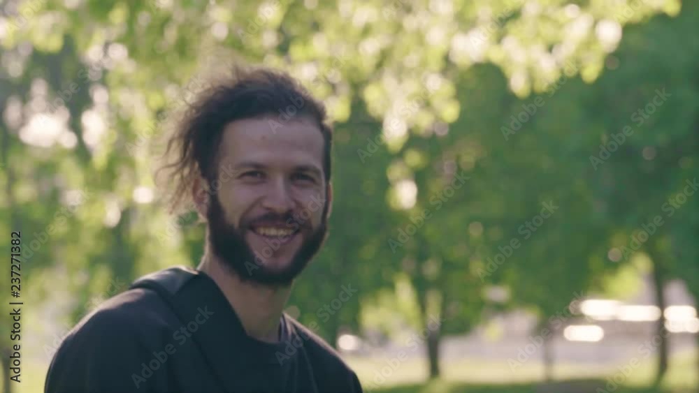 Portrait of a young bearded man standing in sun shining summer park, smiling and then turns back. 4K half-turned side view slowmo Portrait of a young bearded man standing in sun shining summer park, smiling and then turns back. 4K half-turned side view slowmo