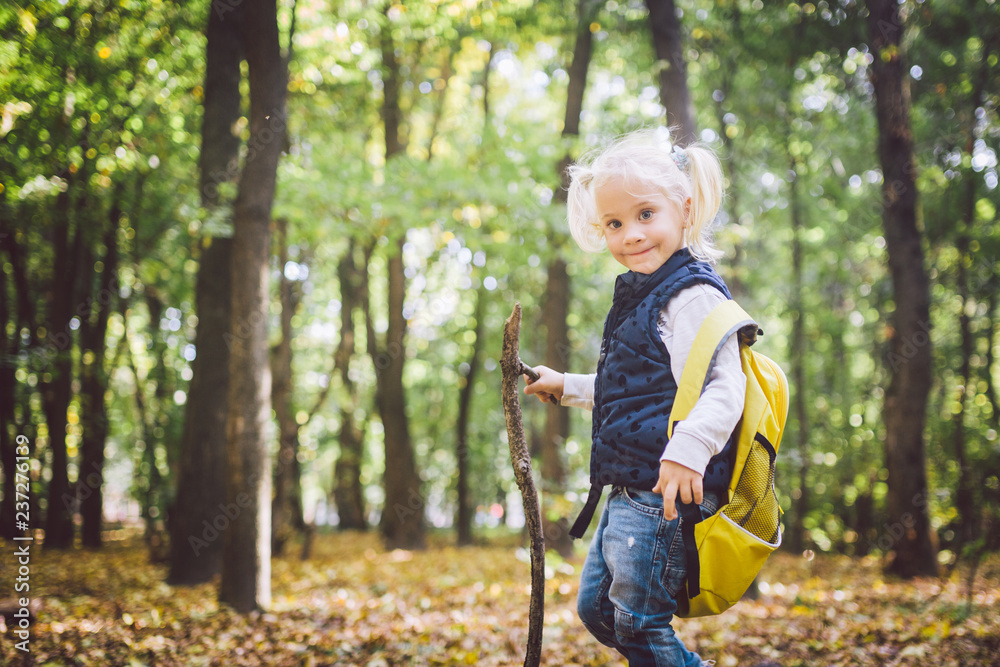 Theme outdoor activities in nature. Funny little Caucasian blonde girl walks walks hiking in the forest on rough terrain with a large backpack. Uses walking stick Theme outdoor activities in nature. Funny little Caucasian blonde girl walks walks hiking in the forest on rough terrain with a large backpack. Uses walking stick