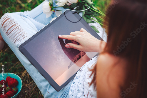 Fototapeta Naklejka Na Ścianę i Meble -  Young woman using a tablet pc sitting in summer grass with bouquet of peonies flowers. Summer vacation