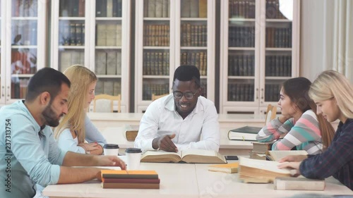 Young african american guy, Arabian man and three caucasian beautiful girls with long blonde hair sitting at table, reading big book in library. photo