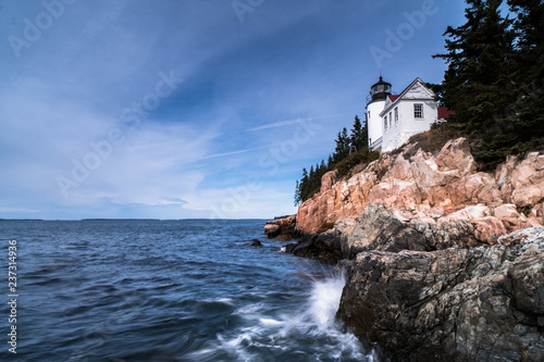 white lighthouse over a blue ocean and red rocks with forest on the rocky coast of Maine under a blue sky