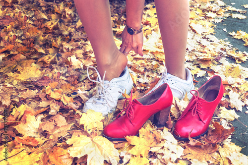 Young woman putting on white sports shoes for active walking in park. Legs of single girl in run snickers on the road among falling yellow maple leaves. Red woman boots wiht high heels. Autumn day.