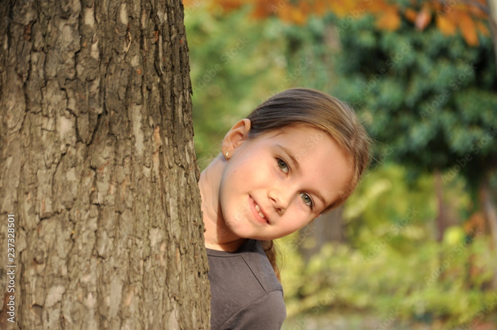 Caucasian white 8 years old child, girl, happy in the park, smiling and hiding behind a big tree Caucasian white 8 years old child, girl, happy in the park, smiling and hiding behind a big tree