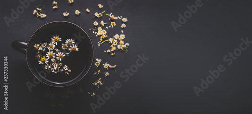 Mug of chamomile tea on dark background  top view with copy space for your design. Healthy tea ingredients and  home remedy for cosmetic treatment. Herbal medicine concept. Template or banner.