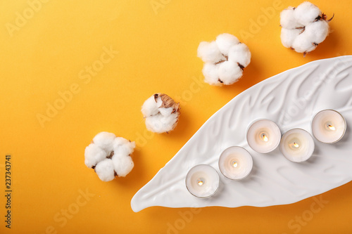 Cotton flowers and plate with burning candles on color background, top view