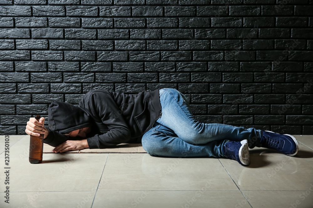 Depressed young man with bottle of beer lying on floor near black brick wall. Alcoholism concept Depressed young man with bottle of beer lying on floor near black brick wall. Alcoholism concept