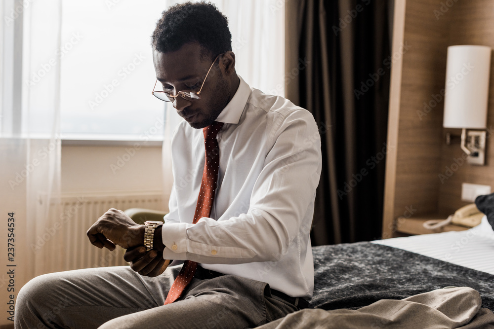 young african american businessman looking at wristwatch while sitting on bed in hotel room