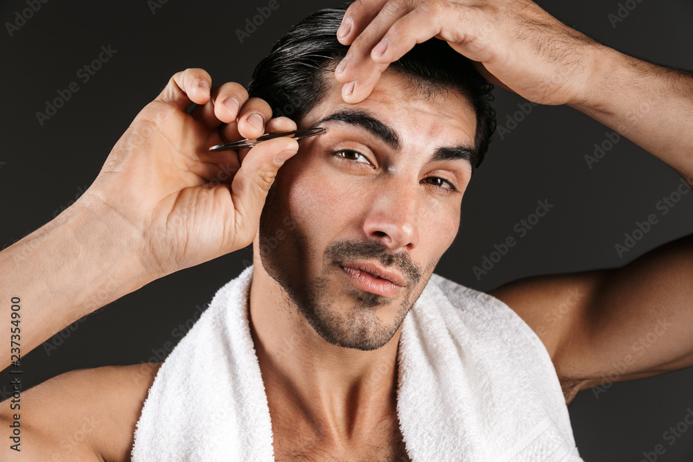 Obraz premium Handsome young man posing isolated over dark wall background correct brows form with tweezer.