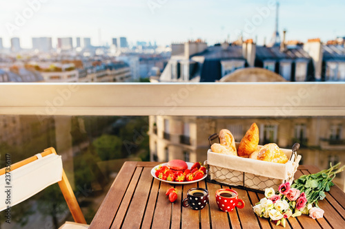 Paris luxury lifestyle. Traditional breakfast. Two cups of coffee espresso, french bakery products, strawberries and flowers on a balcony with a view on parisian rooftops