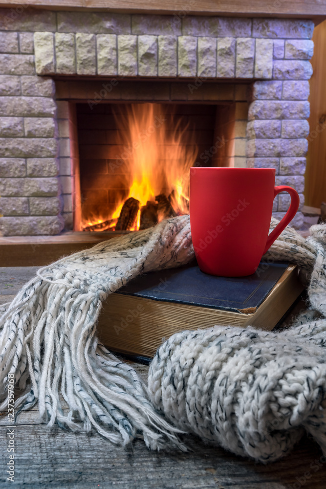 Cozy scene before fireplace with red mug with tea, a book, wool scarf.