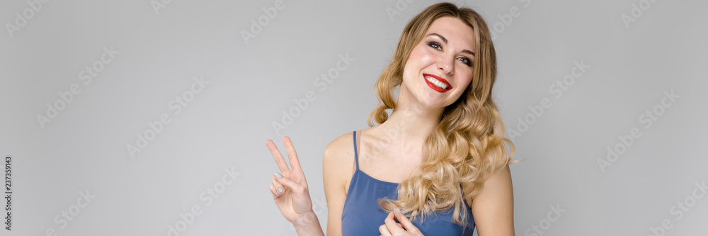 Beautiful girl with strong hair shows two fingers. Girl in a blue top on gray background Beautiful girl with strong hair shows two fingers. Girl in a blue top on gray background