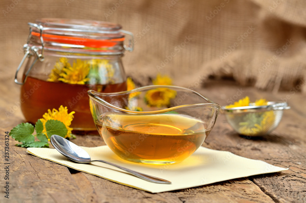 Dandelion jam in glass, spoon, dandelion head around, small colander and full jar of jam in background