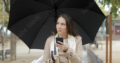 Front view portrait of a frustrated woman checking weather app on a smart phone under the rain in winter