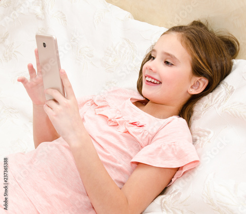 Cute smiling little girl child with her mobile phone smartphone lying on the bed