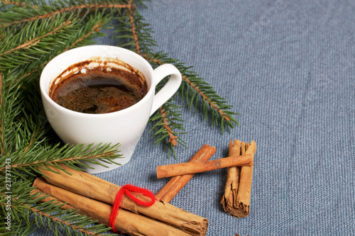 A cup of coffee on the table. Cinnamon sticks next to the fir branches. On a woven background.