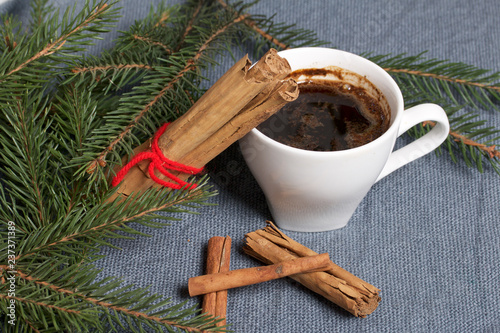 A cup of coffee on the table. Cinnamon sticks next to the fir branches. On a woven background.