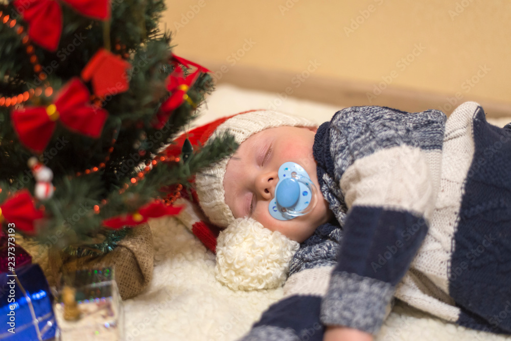 Little baby with nipple in Santa's hat sleeping under the Christmas tree Little baby with nipple in Santa's hat sleeping under the Christmas tree