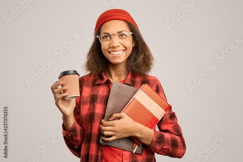 A young smiling woman student in a red plaid shirt and a red hat holds folders with notebooks and documents and a Cup of coffee. Isolated Studio portrait on white background