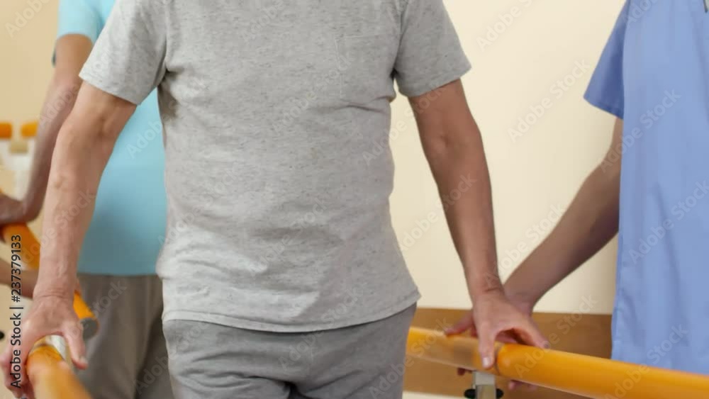 Tilt up shot of elderly man holding on to handrails and learning to walk again after suffering stroke. Male physiotherapist in scrubs talking and supporting him