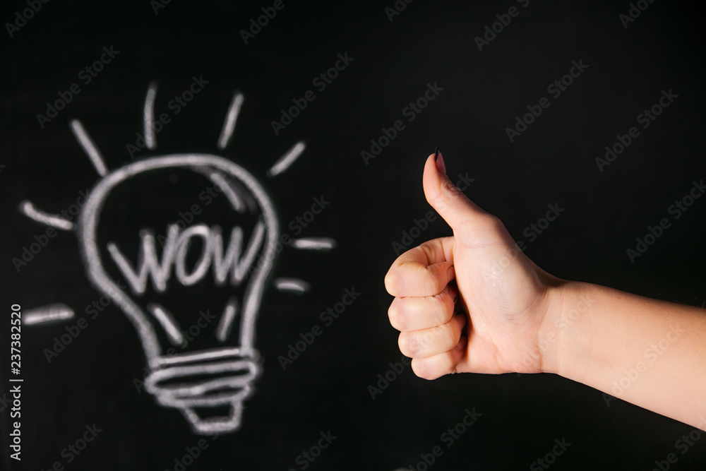 Closeup of female hand with raised thumb near chalked light bulb on black board with word wow Closeup of female hand with raised thumb near chalked light bulb on black board with word wow