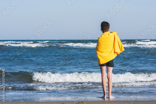 Back view on the boy standing on seashore of the beach in the yellow towel and looking on the sea. Concept.