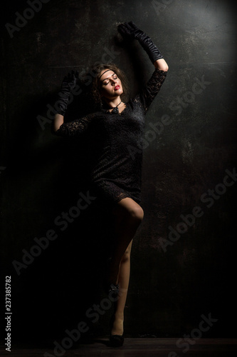 Roaring Twenties. Woman portrait in the style of Gatsby. Low key. Beautiful young woman in a lace black dress, posing sensually, leaning against a black wall.