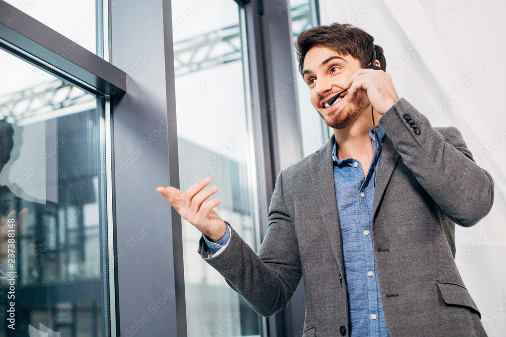 handsome call center operator standing by window, touching headset and speaking in microphone in office handsome call center operator standing by window, touching headset and speaking in microphone in office
