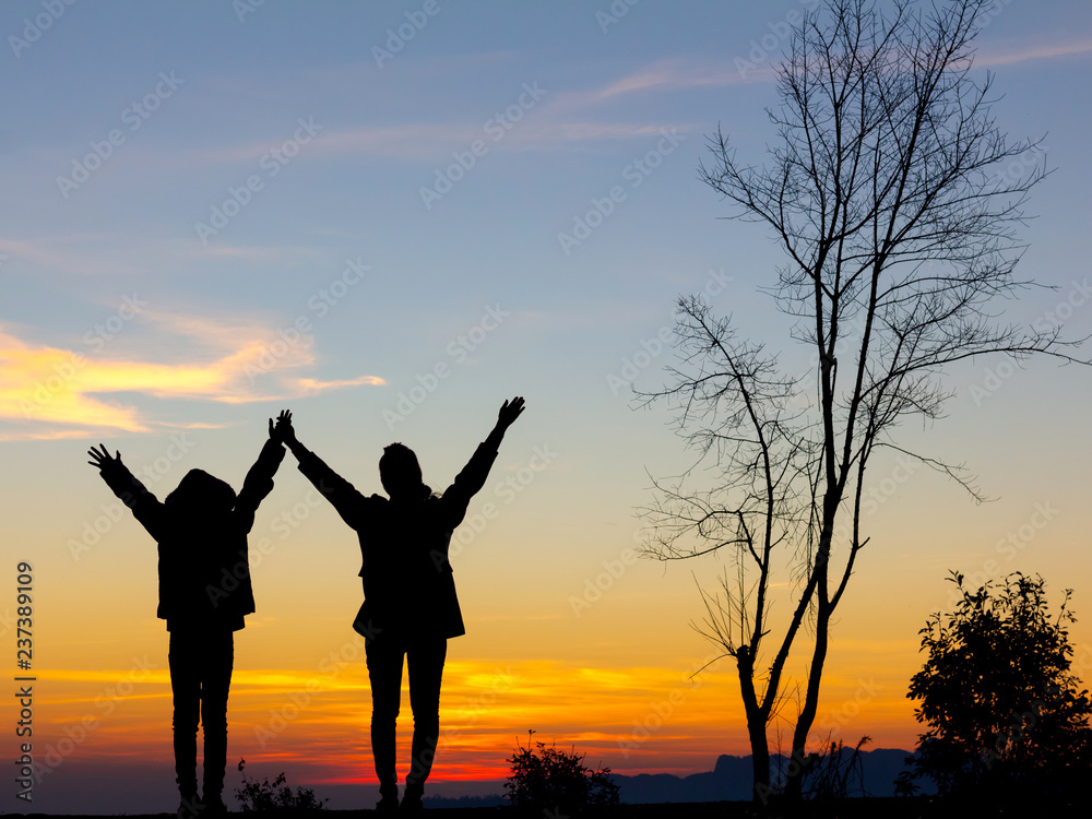 Silhouette of happy two girls standing with raised hands on the mountain at the sunset or sunrise time.