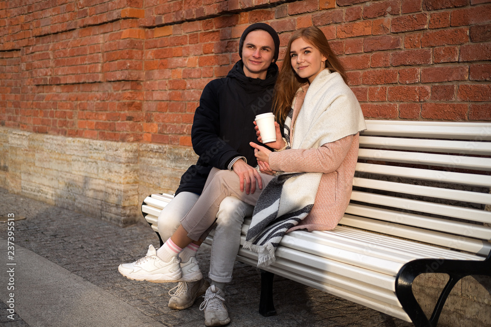 Happy caucasian couple in love in park sitting on the bench near brick wall with takeaway drink Happy caucasian couple in love in park sitting on the bench near brick wall with takeaway drink