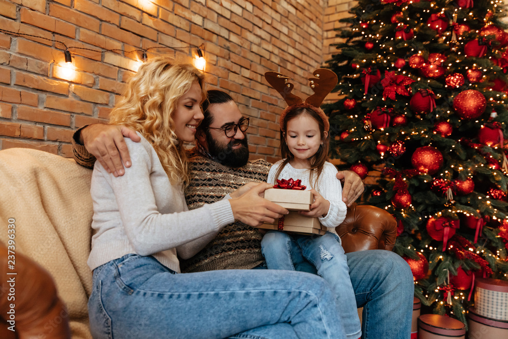 Christmas. Family. Home. Togetherness. Little girl is opening a gift box while sitting with her parents on the couch, all are smiling