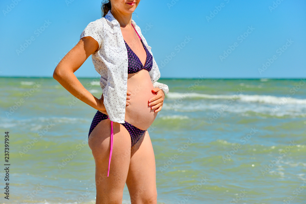 A happy and joyful pregnant woman stands by the sea and ocean on the beach under the sun in the open air, hair develops in the wind. Hands holding on his stomach. A happy and joyful pregnant woman stands by the sea and ocean on the beach under the sun in the open air, hair develops in the wind. Hands holding on his stomach.
