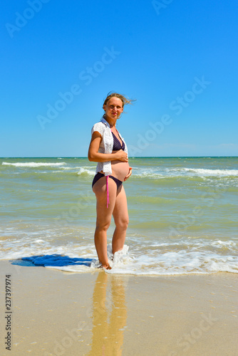 A happy and joyful pregnant woman stands by the sea and ocean on the beach under the sun in the open air  hair develops in the wind.