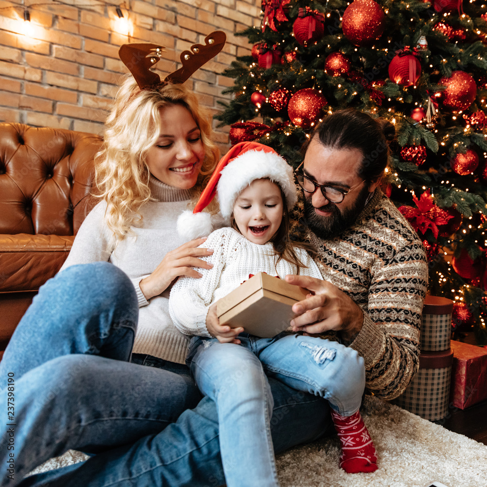 Christmas. Family. Home. Togetherness. Happy parents and their little girl are holding a gift and smiling near the Xmas tree Christmas. Family. Home. Togetherness. Happy parents and their little girl are holding a gift and smiling near the Xmas tree