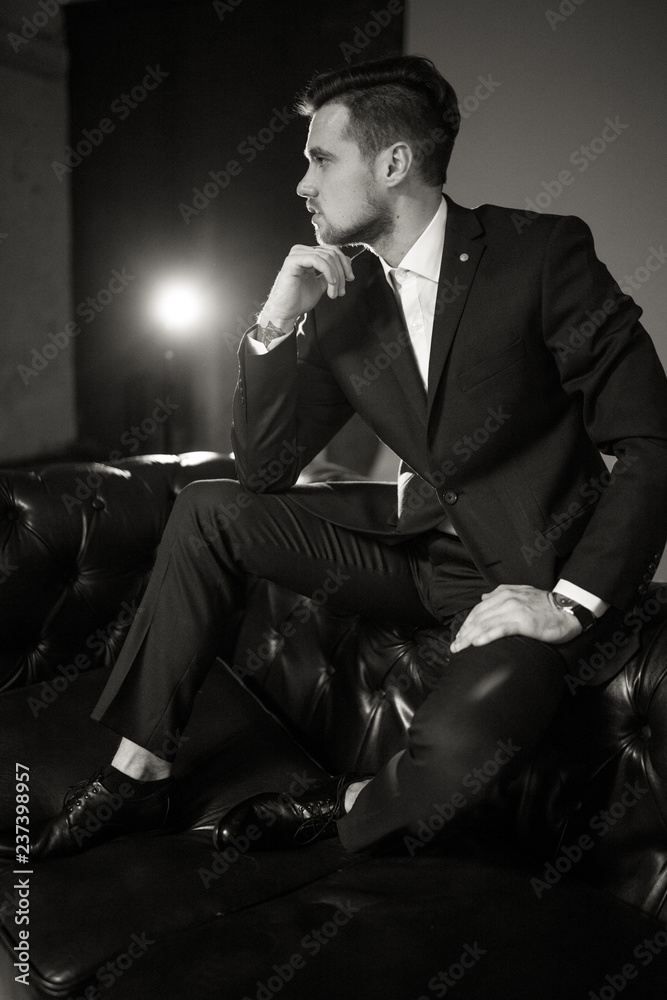 Young businessman in a black suit and a white shirt is sitting and pondering on the leather sofa against a light of lamp. Black and white image. Young businessman in a black suit and a white shirt is sitting and pondering on the leather sofa against a light of lamp. Black and white image.