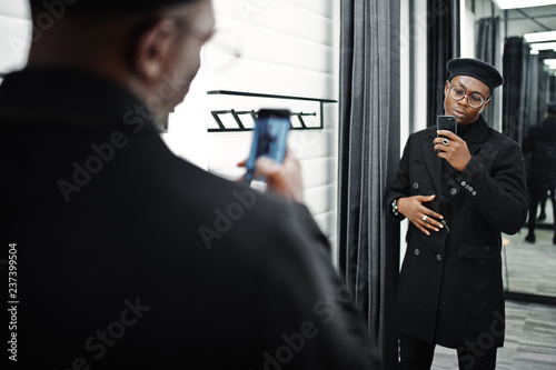 Stylish casual african american man at black beret and overcoat at fitting room clothes store, making photo at mirror on his phone camera. photo