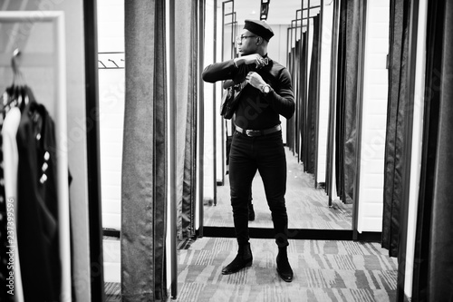 Stylish casual african american man at black outfit and beret with waist bag at fitting room clothes store   looking at mirror.