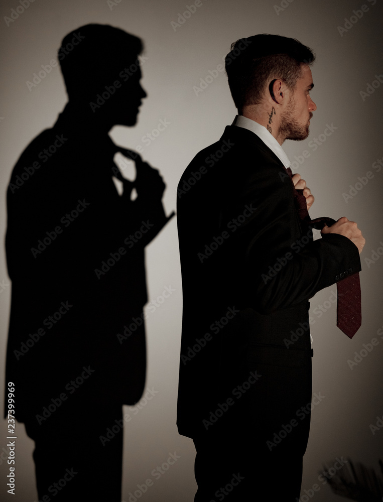 Young businessman in a suit and a shirt is standing against his shadow on the wall.