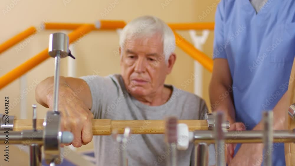 Medium shot with rack focus of elderly man with grey hair using wrist mobility exerciser as unrecognizable male physiotherapist helping and showing the right technique