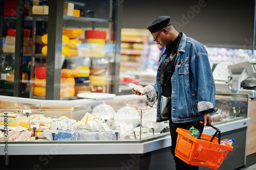 Stylish casual african american man at jeans jacket and black beret holding basket, standing near cheese fridge and shopping at supermarket.