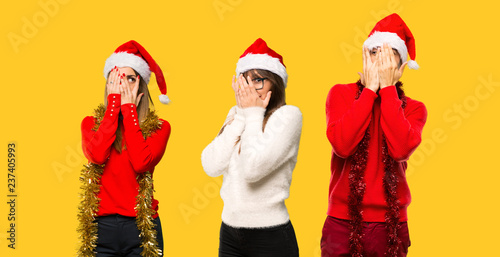 A group of people Blonde woman dressed up for christmas holidays covering eyes by hands and looking through the fingers on yellow background