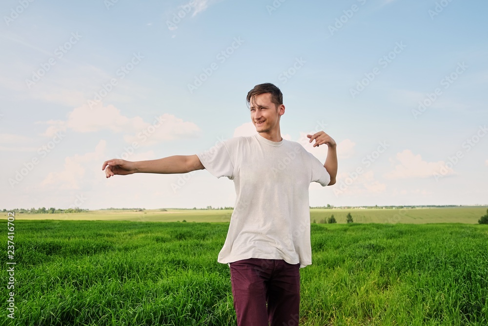 Attractive young man in a white T-shirt, standing in a field of young green wheat, smiling, nature and a sunny summer day with white clouds in the blue sky, throwing his arms to the side