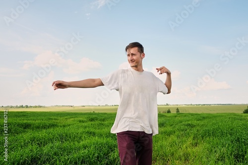 Obraz na płótnie Attractive young man in a white T-shirt, standing in a field of young green wheat, smiling, nature and a sunny summer day with white clouds in the blue sky, throwing his arms to the side w salonie