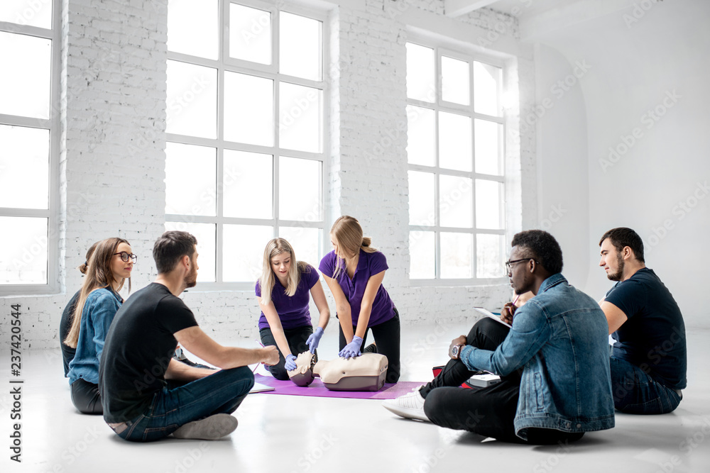 Group of young people sitting in a circle during the first aid courses with two instructors training on medical dummies in the white spacious room Group of young people sitting in a circle during the first aid courses with two instructors training on medical dummies in the white spacious room