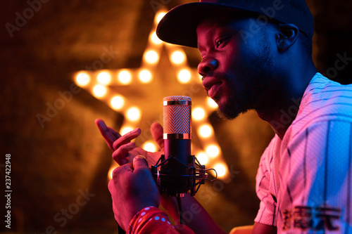 Studio portrait of young man with beard and mustaches in baseball cap and t-shirt with microphone. Pink neon light, star on background