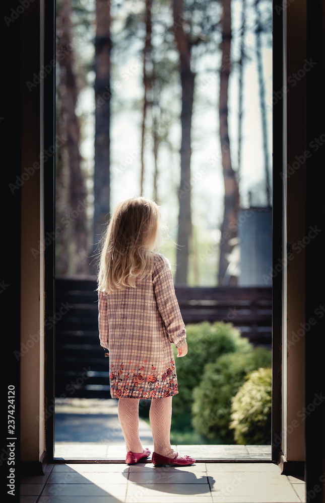 A baby girl with long hair and a beautiful dress looks thoughtfully at the trees in the forest