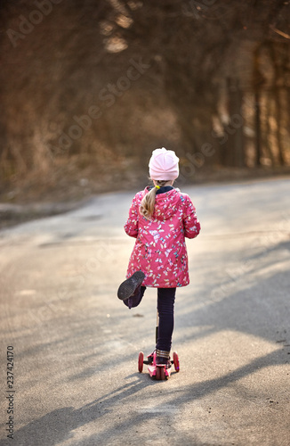Cute little girl in a red jacket riding a scooter down the alley in the park in autumn