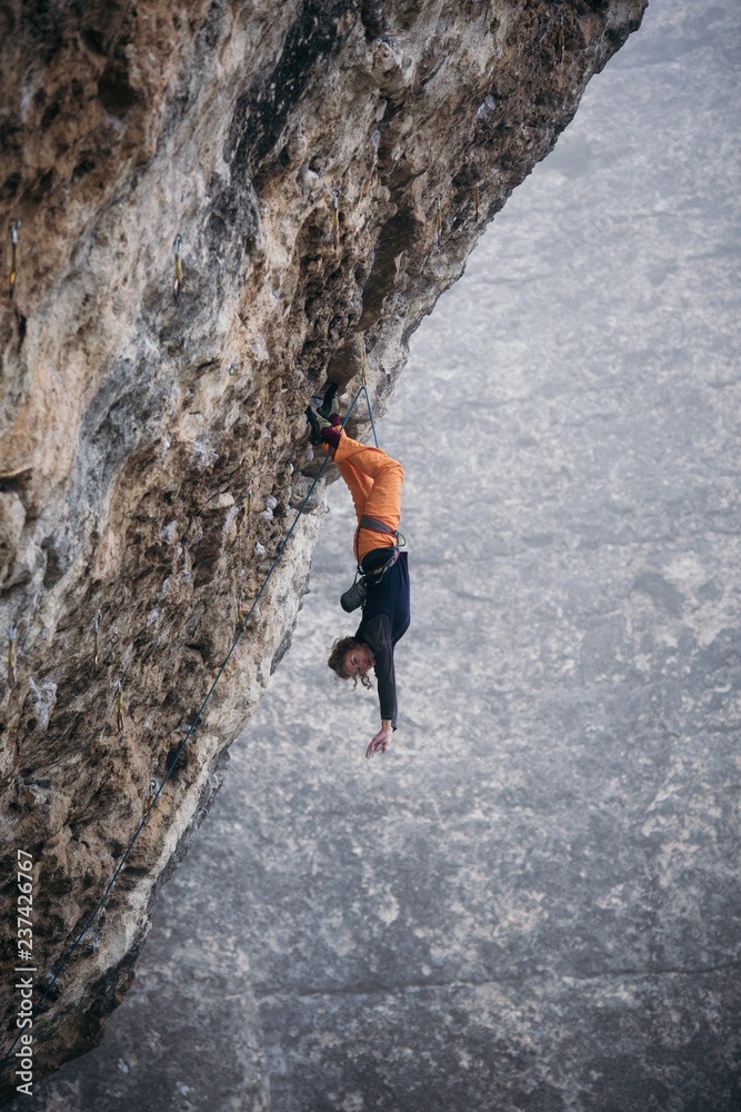 woman-climber hangs upside-down in a grey-color rock in orange pants