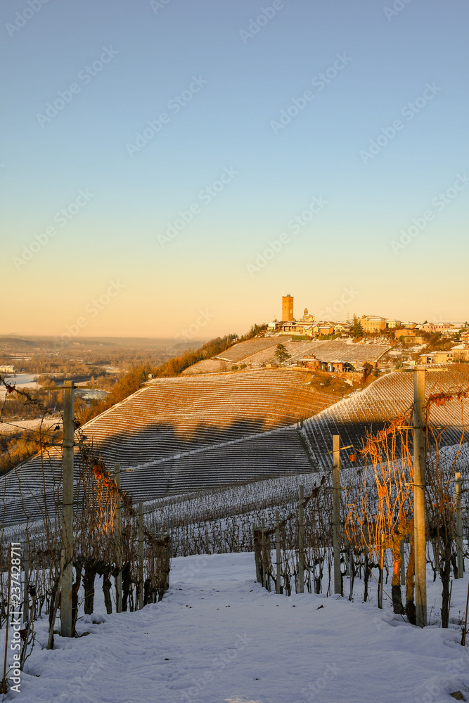 View of snow covered vineyard hills with a small village at the top at sunset in winter, Barbaresco, Langhe, Piedmont, Italy