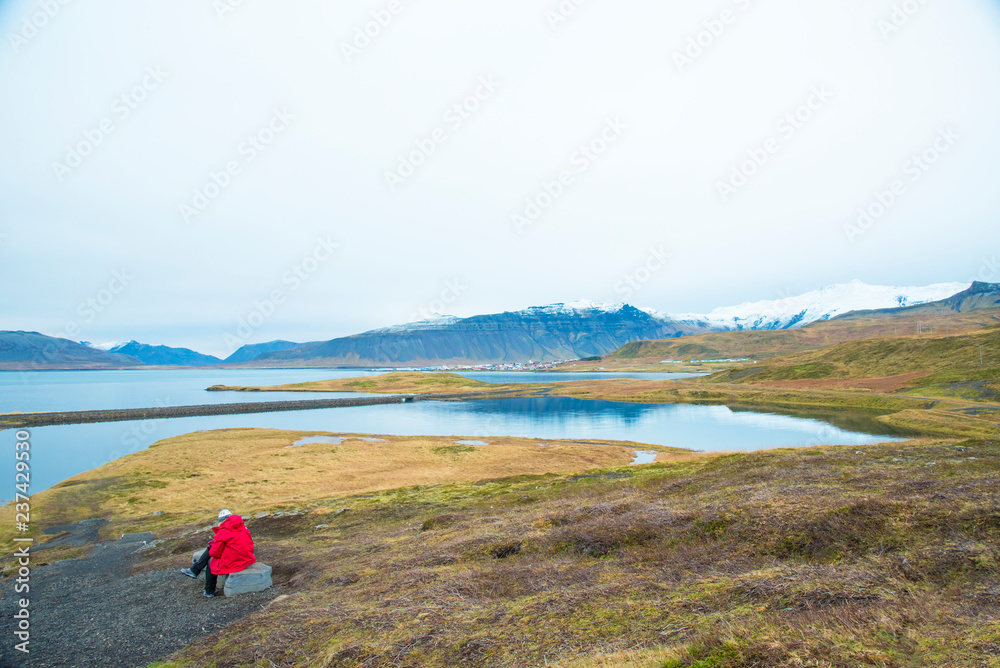 A lady traveller sitting on rocks along the road is looking at the view of mountain range covered by snow in Iceland